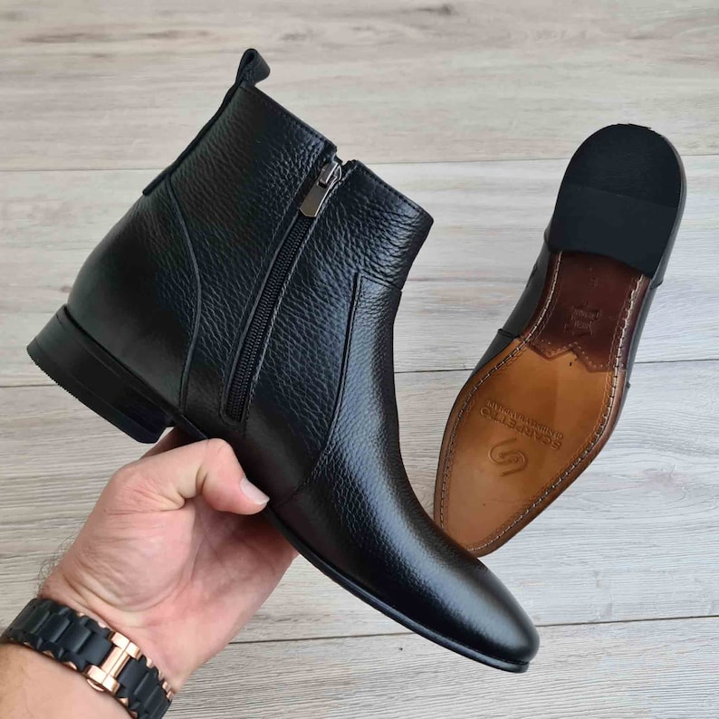 Puede incluir: Botas de tobillo de cuero negro con cremallera lateral y punta puntiaguda. Las botas tienen una suela de cuero marr&oacute;n con el nombre de la marca "SCAMMETTE" impreso en ella.