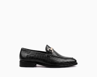 May include: A black loafer shoe featuring a textured crocodile-embossed pattern across the upper. The shoe includes a metallic gold-toned horsebit detail across the vamp and a low, stacked heel, presented from a side profile view.