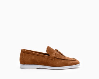 May include: A tan suede loafer featuring a moccasin-style stitched toe, a decorative tassel detail on the vamp, and a flat white rubber sole. The shoe is crafted from a soft, textured material with clean lines and a low-profile silhouette.