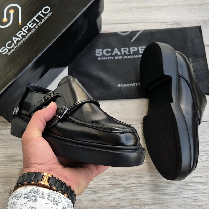 May include: Black leather monk strap shoes with a thick sole. The shoes have two buckles and are displayed with a shoe box and a dust bag, both with the text "SCARPETTO QUALITY AND ELEGANCE AT YOUR REACH".