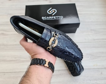 May include: A pair of navy blue leather loafers with a gold buckle and a paisley pattern. The loafers are on a wooden surface.