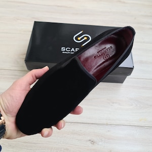 May include: Black velvet loafer with a red leather lining and the text "Scarpetta" in gold lettering. The loafer is on a black box with the text "Scaf" and "Quality Apparel" in white lettering.
