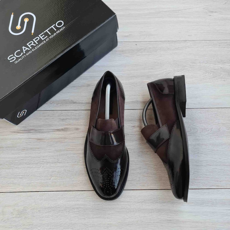 May include: A pair of brown leather slip-on shoes with a black patent leather toe cap and wingtip detailing. The shoes have a brown suede panel on the side and a black leather heel. The shoes are on a light wood surface. The text "SCARPETTO QUALITY AND ELEGANCE AT YOUR REACH" is on a black box in the background.
