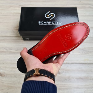 May include: A close-up of the red leather sole of a black shoe. The sole has a stitched edge and the brand name "Scarpetto" is embossed in the leather.
