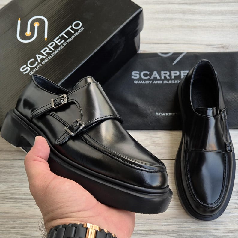 May include: Black leather monk strap shoes with double buckles and a thick sole. The shoes are displayed with a black box and a black dust bag, both bearing the brand name "SCARPETTO". The shoes are a classic style.