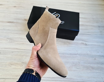 May include: A beige suede ankle boot with a pointed toe and a black sole. The boot is being held by a hand.