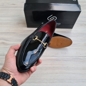 May include: Black leather loafers with a gold metal buckle. The loafers have a shiny finish and a pointed toe.