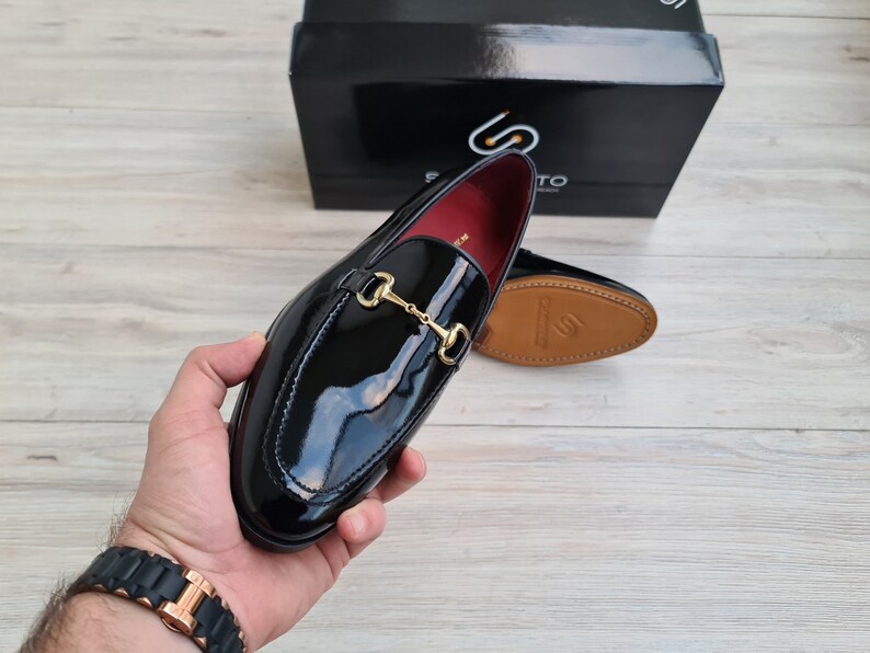 May include: Black leather loafers with a gold metal buckle. The loafers have a shiny finish and a pointed toe.