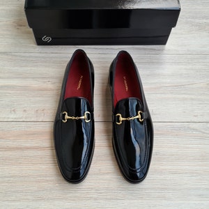 May include: A pair of black patent leather loafers with gold metal buckles. The loafers have a pointed toe and a low heel.
