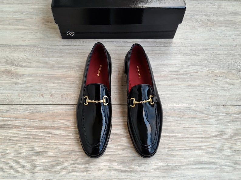 May include: A pair of black patent leather loafers with gold metal buckles. The loafers have a pointed toe and a low heel.