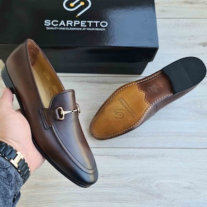 May include: Brown leather loafers with a gold metal buckle detail. The shoes have a leather sole and are made by Scarpetto, a brand that emphasizes quality and elegance.