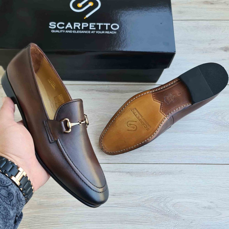 May include: Brown leather loafers with a gold metal buckle detail. The shoes have a leather sole and are made by Scarpetto, a brand that emphasizes quality and elegance.