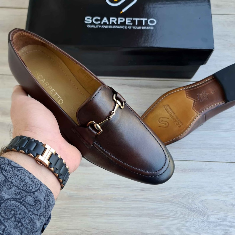 May include: Brown leather loafers with a gold metal buckle detail. The loafers have a leather sole and are made by the brand Scarpetto.