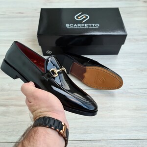 May include: A pair of black leather loafers with a gold buckle detail. The loafers have a pointed toe and a low heel. The shoes are being held by a hand wearing a black watch.