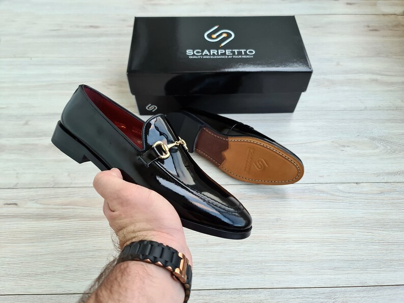 May include: A pair of black leather loafers with a gold buckle detail. The loafers have a pointed toe and a low heel. The shoes are being held by a hand wearing a black watch.