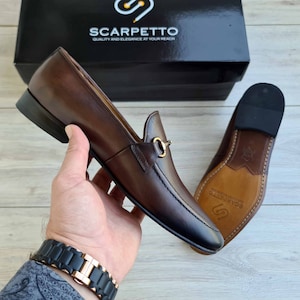 May include: Brown leather loafers with a gold buckle detail. The shoes have a pointed toe and a leather sole. The brand name "Scarpetto" is printed on the sole.