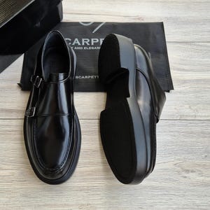 May include: Pair of black leather double monk strap shoes with a thick sole. The shoes have a shiny finish and two silver buckles. The soles are black and the shoes are displayed on a light wood surface.