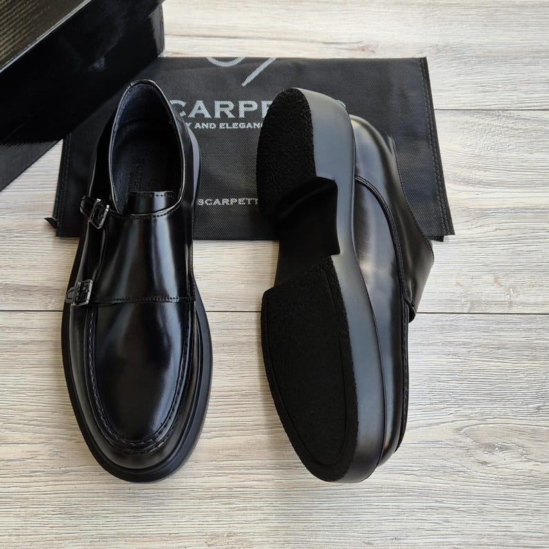 May include: Pair of black leather double monk strap shoes with a thick sole. The shoes have a shiny finish and two silver buckles. The soles are black and the shoes are displayed on a light wood surface.