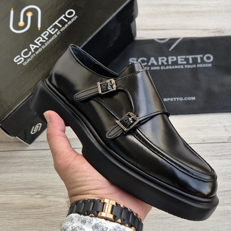 May include: Black leather monk strap shoes with silver buckles and a thick sole. The shoes are displayed with a box and a bag, both with the text "SCARPETTO QUALITY AND ELEGANCE AT YOUR REACH".