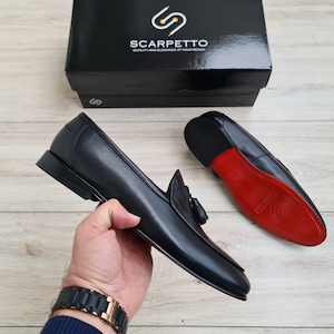 May include: A pair of black leather tassel loafers with red leather soles. The loafers are on a wooden surface. The brand name "Scarpetto" is visible on a black box in the background.