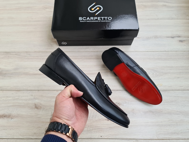 May include: A pair of black leather tassel loafers with red leather soles. The loafers are on a wooden surface. The brand name "Scarpetto" is visible on a black box in the background.