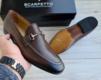Könnte beinhalten: Braune Lederschuhe mit goldener Metallschnalle. Die Schuhe haben eine spitze Zehenpartie und eine Ledersohle. Der Markenname "Scarpetto" ist auf der Sohle aufgedruckt.