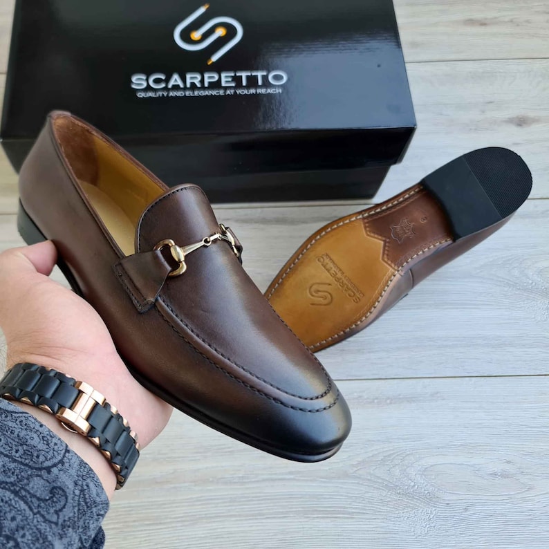 May include: Brown leather loafers with a gold metal buckle detail. The loafers have a pointed toe and a leather sole. The brand name "Scarpetto" is printed on the sole.