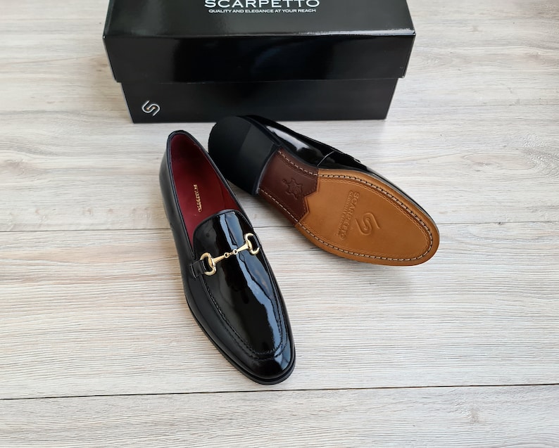 May include: A pair of black patent leather loafers with gold metal buckles. The loafers have a brown leather sole and are made by the brand Scarpetto.