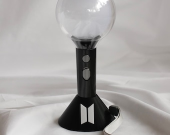 May include: A black and clear light stick with a round head and a black base. The light stick has two silver buttons and a white logo on the base. A gray strap with "BTS" is attached.