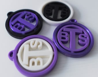 May include: Four round keychains with the letters "BTS" in different colors and styles. Two keychains are purple with white letters, one is black with white letters, and one is white with black letters.