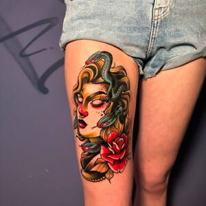 May include: A colorful tattoo of Medusa on a person's thigh. The design features Medusa's face with snakes for hair, a red rose, and a pearl necklace. The tattoo is done in a traditional style with bold lines and vibrant colors.