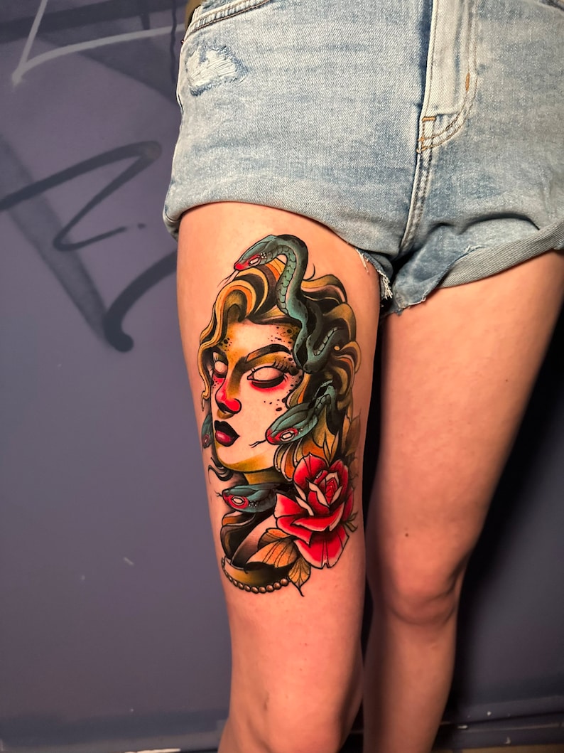 May include: A colorful tattoo of Medusa on a person's thigh. The design features Medusa's face with snakes for hair, a red rose, and a pearl necklace. The tattoo is done in a traditional style with bold lines and vibrant colors.