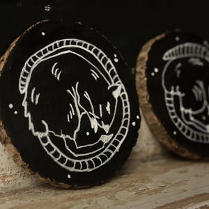 May include: Two black wooden coasters with white painted designs of sleeping animals. The coasters are round and have a natural wood edge.
