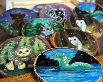 May include: A collection of eight hand-painted wooden round ornaments featuring various mythical creatures, including a sasquatch, a deer, a moth, a loch ness monster, and an owl. The ornaments are painted in a whimsical style with bright colors and a starry night sky background.