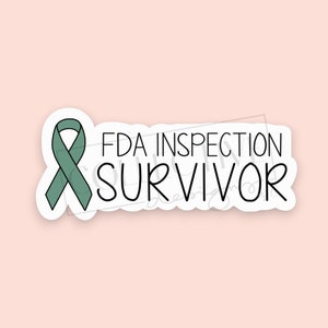 May include: A white sticker with the text "FDA INSPECTION SURVIVOR" in black, accompanied by a teal ribbon. The sticker is on a light pink background. The design is simple and bold, suitable for various applications.