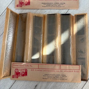 Può includere: Un portafoglio vintage per fotocamera Contax contenente sei strisce di negativi 35mm sviluppati. Il portafoglio in carta color marrone chiaro presenta testo rosso e un'illustrazione di una fotocamera Zeiss Ikon, citando Norman Willet's Camera Center.