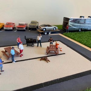 May include: A miniature scene with a motorhome, cars, and people gathered around a picnic table and a grill. The scene is set on a black and white surface with green grass in the background.
