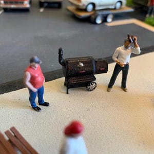 Könnte beinhalten: Miniatur-Grill in Schwarz mit Schornstein, Rädern und einem seitlichen Smoker. Zwei Miniaturfiguren stehen in der Nähe, eine in rotem Hemd und blauer Jeans, die andere in weißem Hemd und schwarzer Hose. Eine Picknick-Szene.