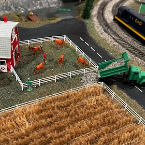Pode incluir: Uma cena de fazenda em miniatura com um celeiro vermelho, uma cerca branca e um campo de feno marrom. Há cavalos em miniatura e pessoas na cena. Um caminhão basculante verde está descarregando cascalho perto do campo.