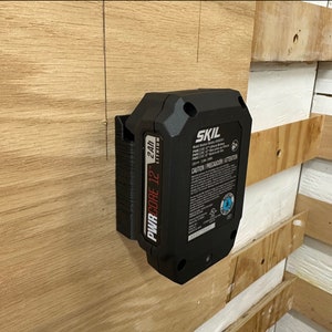 Könnte beinhalten: Ein schwarzer SKIL PWRCORE 12 24V Lithium-Akku, der an einer Holzwand montiert ist. Der Akku hat ein Etikett mit dem Text "CAUTION/PRECAUCION/ATTENTION".