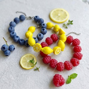 Puede incluir: Tres pulseras de cuentas con dijes de frutas realistas. Una pulsera tiene cuentas de arándanos azules, otra de limones amarillos con rodajas y otra de frambuesas rojas. Cada pulsera incluye una cadena extensora de metal en tono plateado.