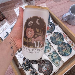 May include: A white glass can-shaped tumbler with a bamboo lid. The tumbler features a brown graphic of flowers, mushrooms, a moon, and butterflies, with the text "AND IF NOT, HE IS STILL GOOD - Daniel 3:18". Car coasters with various designs are also visible.