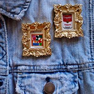 May include: Two gold-framed pins on a blue denim jacket. The left pin features a colorful abstract painting with the word "MATISSE" above it. The right pin features a red and white can of Campbell's Tomato Soup.