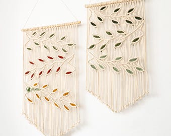 May include: Two macrame wall hangings with leaf designs in shades of green, red, and yellow. The wall hangings are cream-colored with a wooden dowel at the top and fringe at the bottom. They are hanging on a white wall above a wooden cabinet.