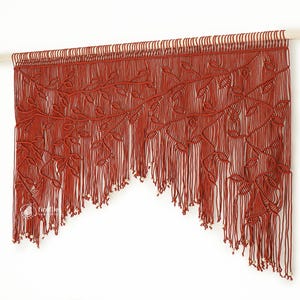 Pode incluir: Uma tapeçaria de parede em macramé cor de ferrugem com um design botânico. A peça feita à mão apresenta nós intrincados que formam folhas e vinhas, com uma borda inferior com franjas. É montada em um pino de madeira, adicionando um toque natural.