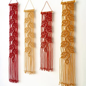 May include: Four macrame wall hangings in shades of red, tan, and gold, each featuring a leaf design. The hangings are suspended from triangular frames and are displayed against a white wall, above a red sofa with decorative pillows.