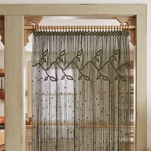 May include: A beaded curtain hangs in a doorway, featuring a dark green color with leaf-like designs. The curtain is suspended from a wooden rod. The room has a natural, earthy aesthetic, with a jute rug and wooden furniture.