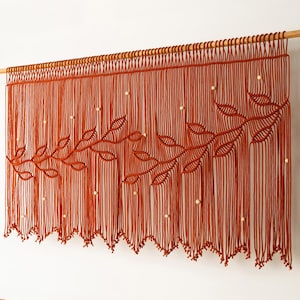 May include: A rust-colored macrame wall hanging with a wooden dowel. The design features a leaf pattern and small, round, white accents. The macrame is hanging above an orange sofa in a living room setting.