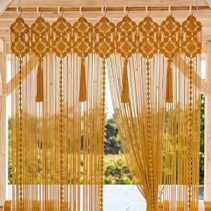 May include: A mustard yellow macrame curtain featuring an intricate knotted diamond pattern at the top, long vertical fringe strands, and decorative tassels hanging from a wooden rod, creating a textured divider with a repeating geometric design.