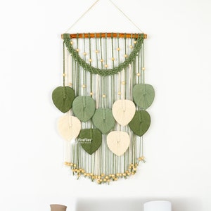 May include: A green and cream macrame wall hanging with wooden beads and leaf shapes. The hanging is on a white wall above a wooden table with a vase, a basket, and a framed picture.